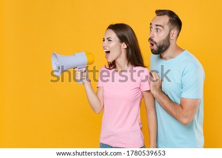Shocked young couple two friends guy girl in blue pink t-shirts posing isolated on yellow wall background studio. People lifestyle concept. Mock up copy space. Screaming in megaphone looking aside