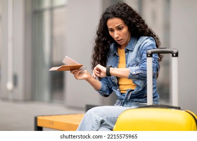 Shocked young curly woman sitting on bench with suitcase, female tourist checking time, looking at smart watch, traveller running late for her flight, waiting for taxi, copy space
