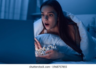 Shocked Young Female Eating Popcorn And Watching Film On Laptop While Resting On Bed In Dark Bedroom At Home