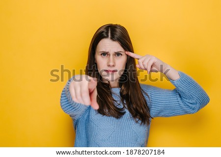 Shocked young female showing stupid gesture and pointing to camera, accusing crazy cuckoo mind, blaming insane idea, reckless plan, dressed in blue sweater, isolated on yellow studio background
