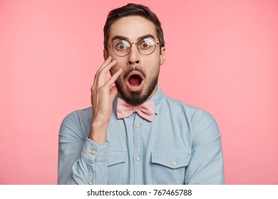 Shocked young handsome man stares through spectacles, being surprised to hear bad news or frightened of future difficuties on work, poses against pink background. Emotional bearded attractive guy