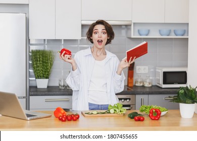 Shocked Young Housewife Woman 20 In White Husband's Shirt And Basic T-shirt Hold Bell Pepper Looking At Recipe In Cookery, Book Preparing Vegetable Salad Cooking Food In Light Kitchen At Home Alone.