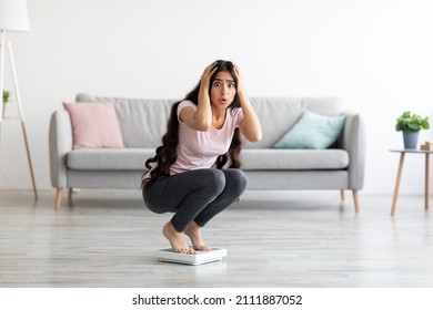 Shocked young Indian lady sitting on scales, terrified of weight gain at home, free space. Millennial woman feeling unhappy with result of her slimming diet. Healthy living concept