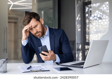 Shocked young man businessman, lawyer, banker sitting in office at desk, holding hand to head and looking frustrated at phone screen. - Powered by Shutterstock