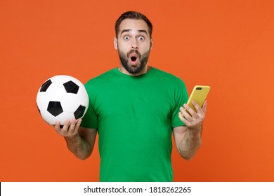 Shocked young man football fan in green t-shirt cheer up support favorite team with soccer ball using mobile cell phone isolated on orange background studio. People sport leisure lifestyle concept - Shutterstock ID 1818262265