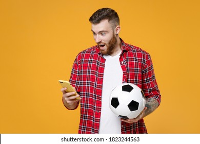 Shocked Young Man Football Fan In Basic Red Checkered Shirt Cheer Up Support Favorite Team With Soccer Ball Using Mobile Cell Phone Isolated On Yellow Background Studio. People Sport Leisure Concept