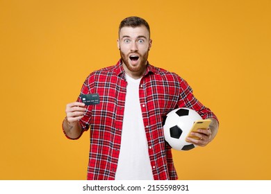 Shocked young man football fan in red shirt cheer up support favorite team with soccer ball using mobile cell phone hold credit bank card isolated on yellow background. People sport leisure concept