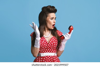 Shocked Young Pinup Woman In Retro Dress Holding Landline Phone Receiver, Feeling Irritated About Telephone Conversation On Blue Studio Background. Unpleasant Contact Concept