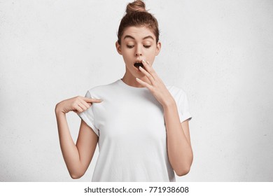 Shocked Young Pretty Female Look Surprisedly Down, Indicats At Blank White T Shirt With Fore Finger, Poses Against Studio Background. Scared Female Finds Out Price On Usual Shirt From Luxury Shop