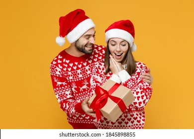 Shocked young Santa couple friends man woman 20s in red sweater Christmas hat hold present box with gift ribbon bow isolated on yellow background. Happy New Year celebration merry holiday concept