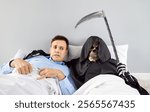 Shocked young scared man lying on bed in bedroom under blanket with skeleton in a grim reaper death costume holding scythe hugging and looking at camera. Halloween and horror concept.