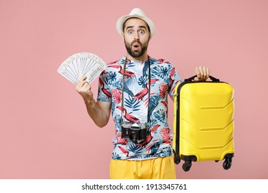 Shocked Young Traveler Tourist Man In Summer Clothes Hat Hold Suitcase Fan Of Cash Money In Dollar Banknotes Isolated On Pink Background. Passenger Traveling On Weekends. Air Flight Journey Concept