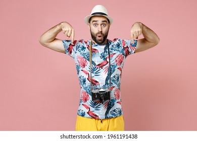 Shocked young traveler tourist man in summer clothes hat with photo camera pointing index fingers down isolated on pink background. Passenger traveling abroad on weekends. Air flight journey concept
