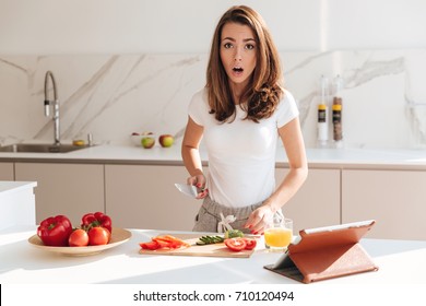 Shocked Young Woman Cooking Salad On A Kitchen With Tablet Computer On A Table