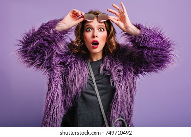 Shocked Young Woman With Curly Blonde Hair Holding Glasses With Mouth Open. Attractive Lady In Purple Fur Coat Posing With Surprised Face Expression.