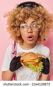 Shocked young woman with curly hair looks amazed away wears broken spectacles safety helmet and gloves eats unhealthy hamburger has quick snack has active journey by bicycle. Junk food concept