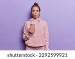 Shocked young woman with hair bun holds modern smartphone reacts to mobile offer dressed in casual pink hoodie stares bugged eyes isolated over purple background checks exciting deal or offer