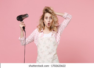 Shocked young woman housewife in casual clothes, apron posing isolated on pastel pink background in studio. Housekeeping concept. Mock up copy space. Blow drying hair with hairdryer, put hand on head