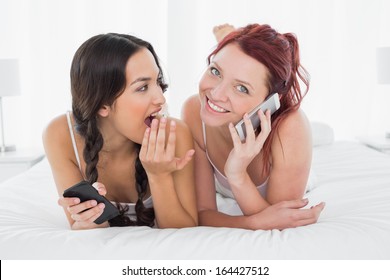 Shocked young woman looking at a cheerful friend while on call in  bed at home