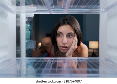 Shocked young woman looking in the empty fridge, she has no food at home, point of view shot from inside the fridge