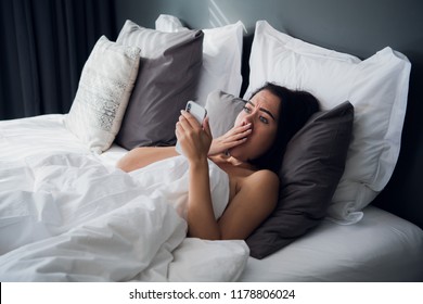 Shocked Young Woman Looking At Mobile Phone In Bed At Home