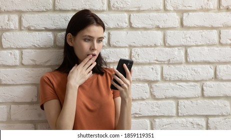 Shocked young woman looking at smartphone screen, getting scam spam social phishing message, reading unbelievable news, feeling frustrated or stressed isolated on brick wall background, copy space.