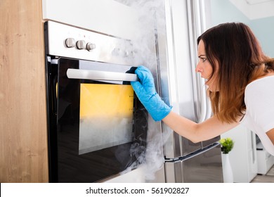 Shocked Young Woman Looking At Smoke Coming Out Of Oven In Kitchen