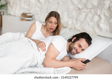 Shocked Young Woman Seeing Her Husband Messaging To His Lover On Smartphone While Lying In Bed At Home. Millennial Wife Feeling Jealous, Finding Out About Her Spouse's Love Affair