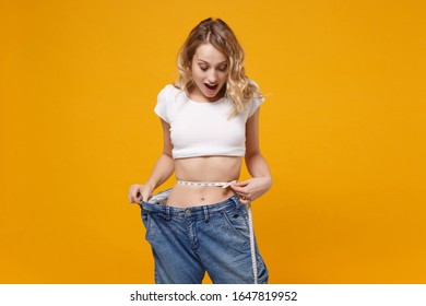 Shocked Young Woman In White T-shirt Isolated On Yellow Orange Background. Proper Nutrition Losing Weight Healthy Lifestyle Dieting Concept. Wearing Old Big Jeans Measuring Waist With Measure Tape