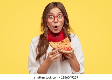 Shocked youngster has snack during dinner time, enjoys delicious pizza, being surprised with its low price, has unhealthy lifestyle, eats junk food, stands against yellow background. Eating concept