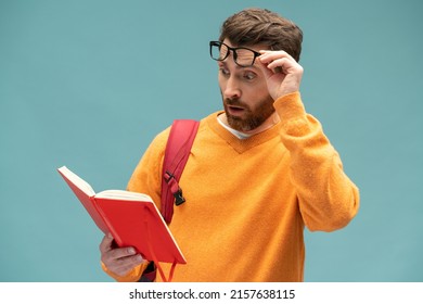 Shocking facts. Astonished smart bearded student man reading book with surprised expression, amazed by story, being shocked by interesting information. Indoor studio shot isolated on blue background 