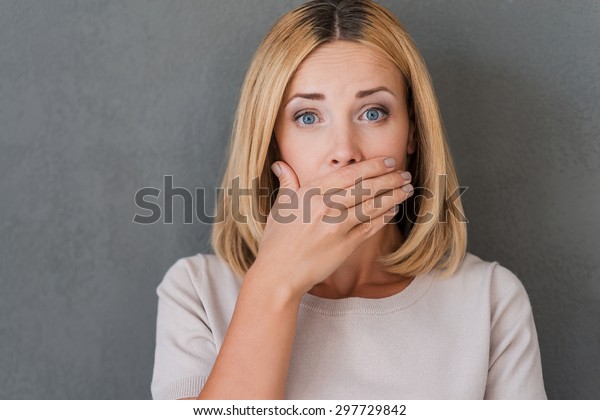 Shocking\
news. Surprised mature woman covering mouth with hand and staring\
at camera while standing against grey\
background