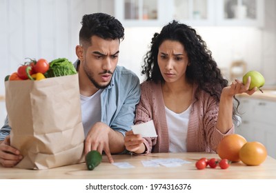 Shocking Prices. Confused Middle Eastern Spouses Checking Bills After Grocery Shopping, Young Arab Couple Sitting At Table In Kitchen, Upset Man And Woman Reading Supermarket Invoice, Closeup