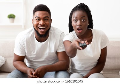 Shocking tv programm. Surprised black couple watching tv with opened mouth, switching channels at camera with dazed face expression
