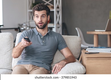 Shocking TV Show. Handsome Young Man Holding Remote Control And Looking Surprised While Watching TV On The Couch At Home