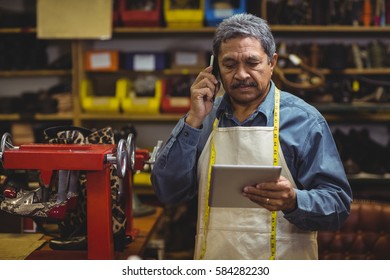 Shoemaker using digital tablet while talking on mobile phone in workshop - Powered by Shutterstock