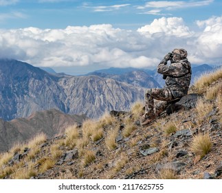 Shooter In Mountain Gear On The Slope Is Watching The Situation Through Binoculars. Hunter Looks Through Binoculars Against The Background Of The Sky And The Mountain Landscape. 