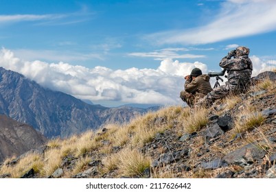 Shooters in mountain gear on the slope are watching the situation through binoculars. Two people are looking through binoculars against the background of the sky and the mountain landscape. 