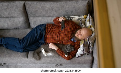 Shooting from above, man lies on couch half-side with cat under his arm, scratches his pet and watches movie on phone. Caucasian man in plaid red shirt is resting on sofa with a cat and a smartphone. - Shutterstock ID 2236348509
