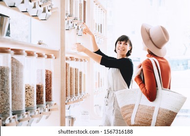 Shop Assistant Helping Customer In Bulk Food Store. Seller Advising Woman In Her Purchase Of Groceries Without Plastic Packaging In Zero Waste Shop. Sustainable Shopping At Small Local Businesses.