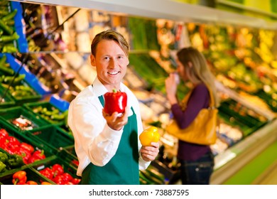 Shop assistant in a supermarket at the vegetable shelf; in the background a woman choosing vegetables - Powered by Shutterstock