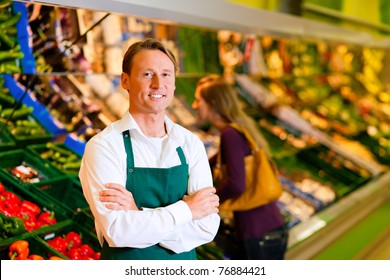 Shop Assistant In A Supermarket At The Vegetable Shelf; In The Background A Woman Choosing Vegetables