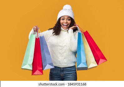 Shopaholic Concept. Portrait Of Excited Black Woman Wearing Winter Hat And Sweater Carrying Bright Colorful Shopping Paper Bags Standing At Studio Isolated Over Orange Background Wall. Sale And Retail