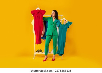 Shopaholic. Happy elegant woman choosing dresses holding two hangers, standing near clothing rail with trendy clothes over yellow background. Female fashion choice