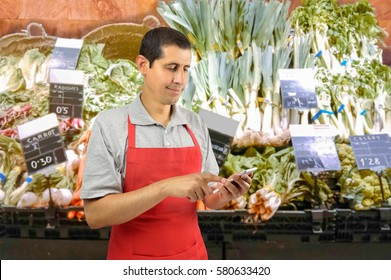 Shopman With Apron Uses A Smart Phone At The Greengrocer