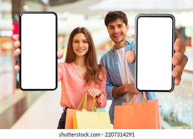 Shopping App. Couple Showing Huge Mobile Phones With Blank Screens Standing Holding Shopper Bags In Modern Hypermarket. Spouses Recommending Application For Customers. Mockup