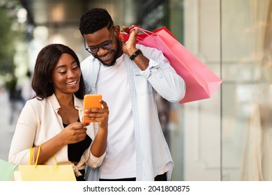 Shopping App. Portrait Of Cheerful African American Spouses Using Smartphone Searching Sales And Special Offers Standing Outdoors Near Mall Store. Smiling Couple Reading Message With Sale Offer