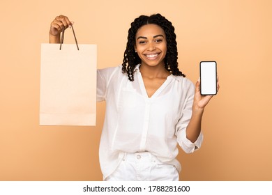 Shopping Application. African American Girl Showing Phone Empty Screen And Shopper Bag Standing On Beige Studio Background. Mockup