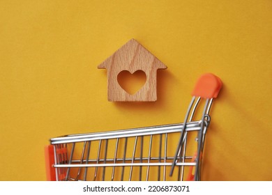 A Shopping Basket And A Wooden House Above It. The Symbol Of Buying A Home