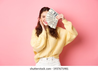 Shopping. Beautiful Brunette Woman Holding Money On Face And Smiling, Touching Cheek, Standing Against Pink Background